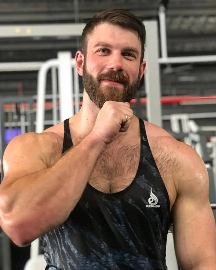 Homme barbu, torse nu, portant un débardeur noir, faisant un geste de poing devant sa bouche, souriant, dans une salle de sport.