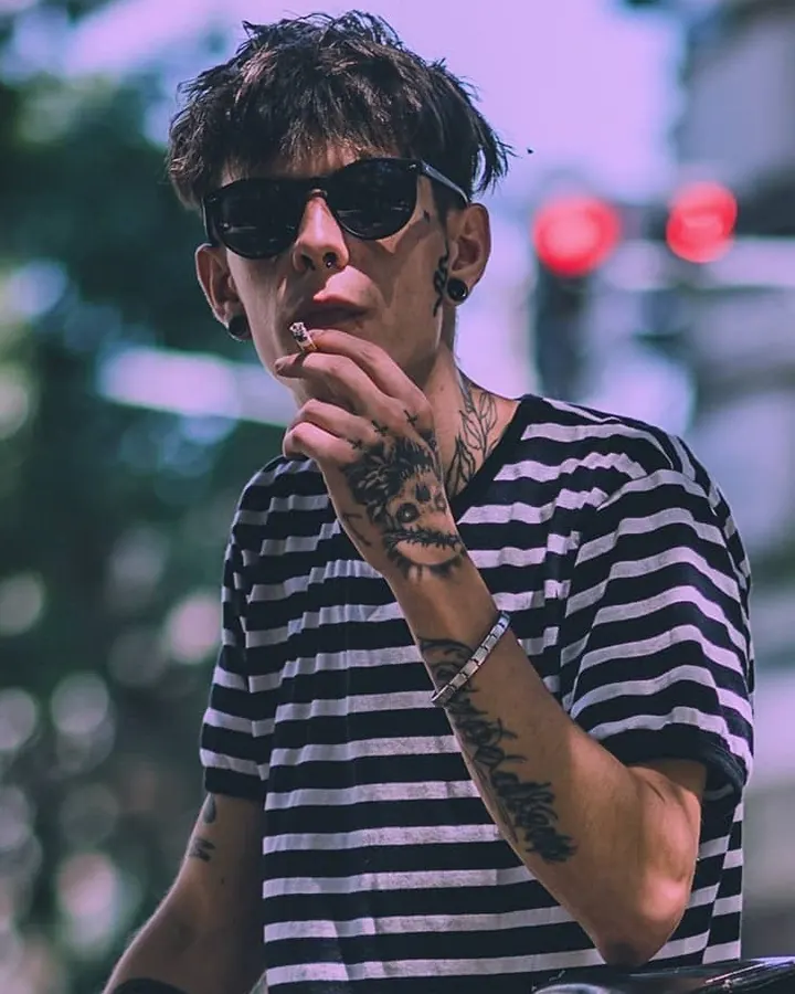 Homme aux cheveux bruns-noirs portant des lunettes de soleil et une chemise rayée, tenant une cigarette, tatouages visibles sur la main et le coude.