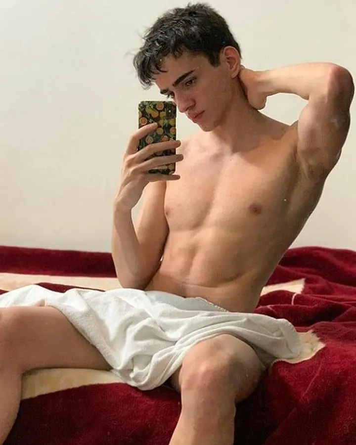 Jeune homme torse nu, portant un short noir, assis sur un lit rouge, tenant un téléphone avec une coque colorée, main derrière la tête, regard vers le bas.