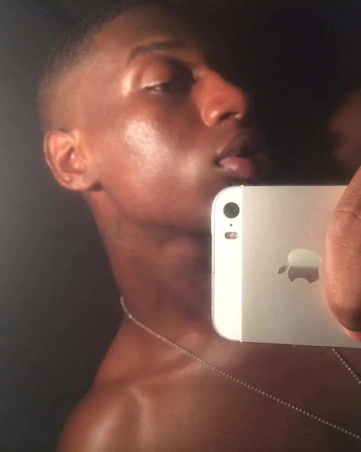 Homme noir torse nu, regard intense, tenant un téléphone argenté, chaîne fine au cou, lumière douce sur la peau.