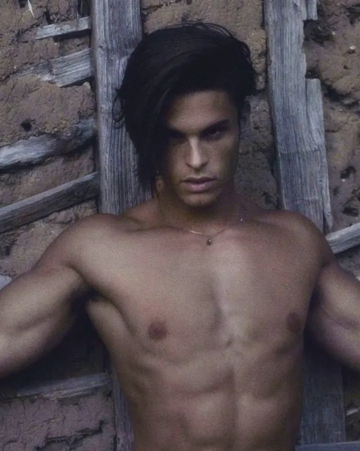Homme européen torse nu, cheveux longs, appuyé contre un mur en bois, regard direct, collier fin, posture détendue mais assurée.