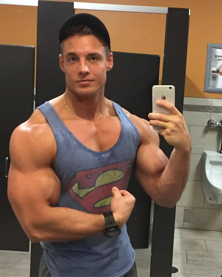 Homme en tank top Superman bleu, casquette noire, prenant un selfie dans un miroir de toilette.