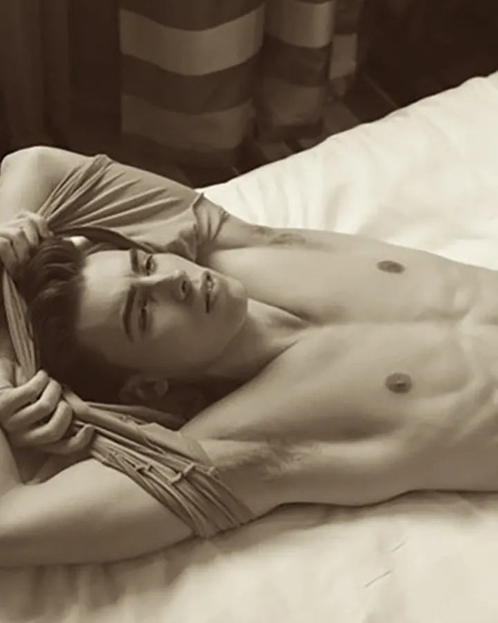 Homme torse nu, barbe courte, regard intense, en sépia, allongé sur un lit, mains derrière la tête.