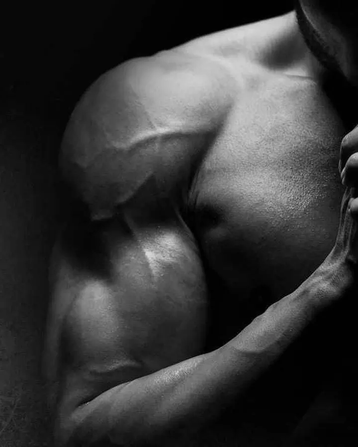 Plan rapproché noir et blanc du torse musclé d’un homme, en lumière dramatique, main posée sur son épaule.