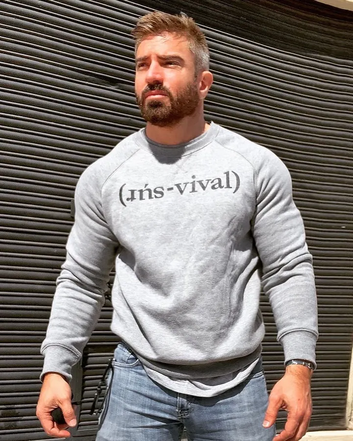 Homme barbu, en sweat gris, debout devant un rideau métallique, regardant vers le côté.