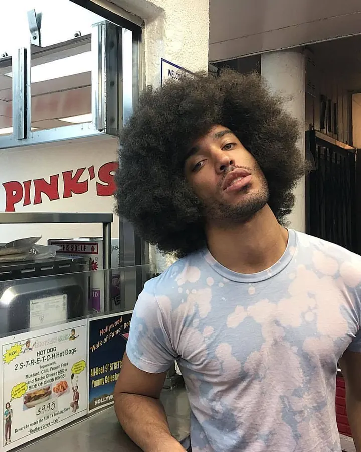 Homme en t-shirt tie-dye bleu et blanc, afro volumineux, regardant l’appareil devant un comptoir de restauration rapide