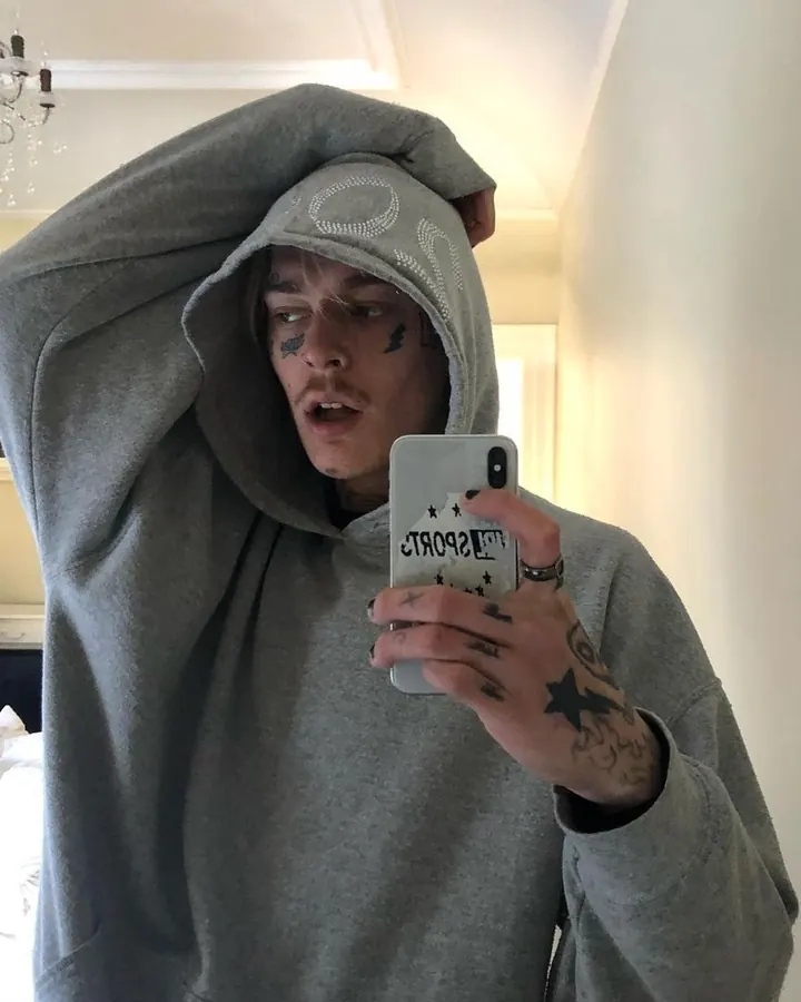 Homme en sweat gris à capuche, prenant un selfie, tatouages visibles sur le visage et la main