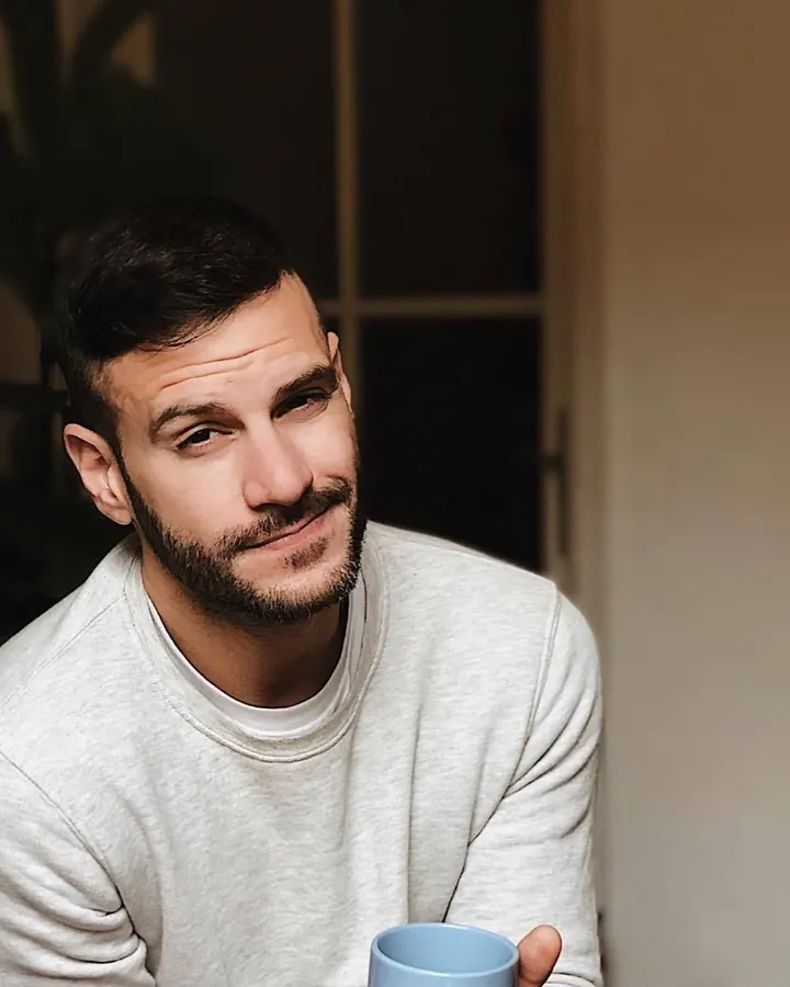 Homme barbu, regardant vers le côté, portant une chemise blanche légèrement ouverte, devant une fenêtre.