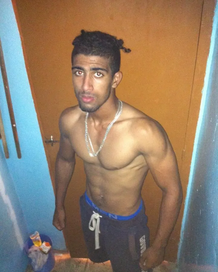 Homme arabe aux cheveux bruns-noirs et barbe, torse nu, portant un sweat gris, devant un mur en bois, en plan moyen.