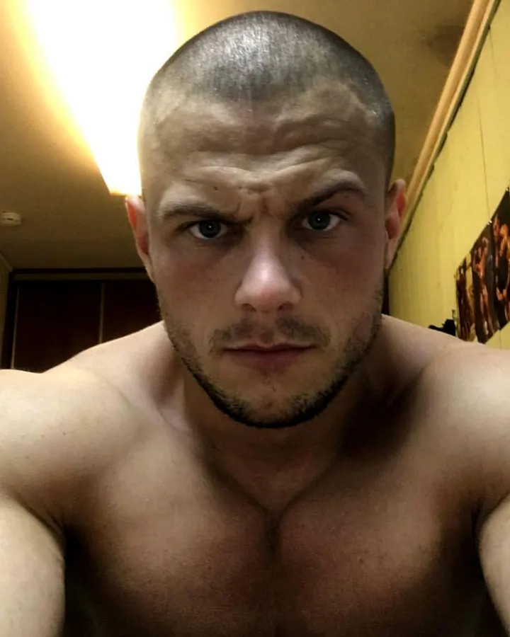 Homme latino torse nu, musclé, en sous-vêtements noirs, assis sur une chaise en face d'un miroir, touchant le cadre.