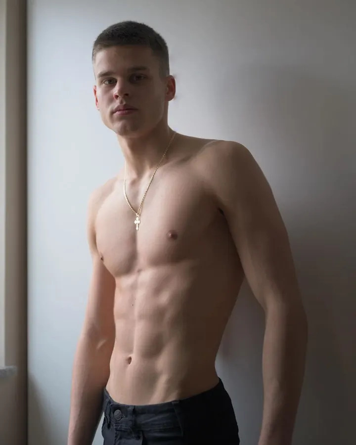 Jeune homme torse nu, debout, regardant directement l'appareil photo, avec une chaîne dorée autour du cou.