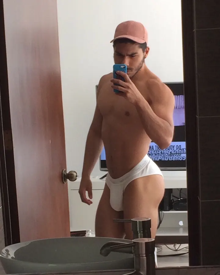 Homme torse nu, portant une casquette rose, prenant un selfie devant un miroir dans une salle de bain, en sous-vêtements blancs.